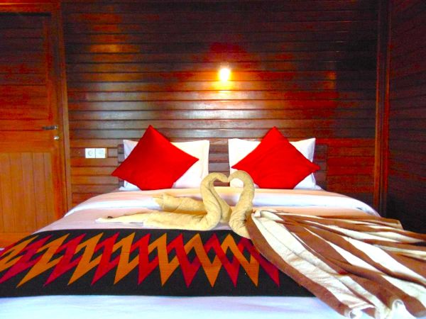 Cozy hotel bed in wooden room with red pillows, towel swans, and colorful woven bedspread. - Bali Villas
